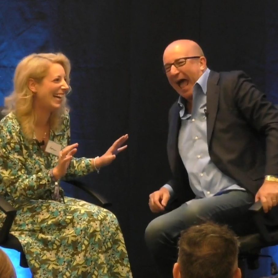 Rebecca, in a patterned dress, and Paul McKenna in a blazer engage in conversation on stage.