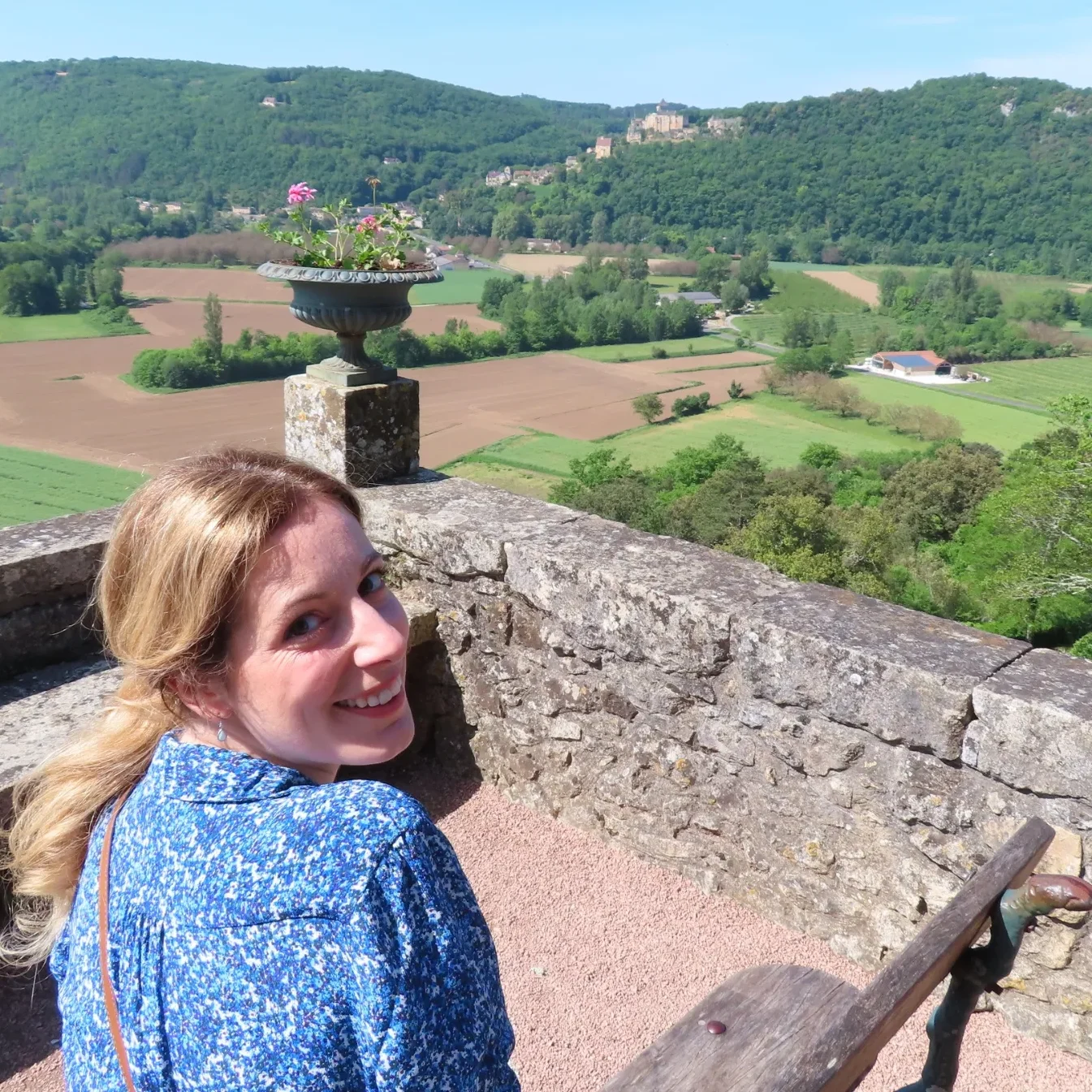 A smiling woman sits on a stone terrace overlooking lush green fields and hills.