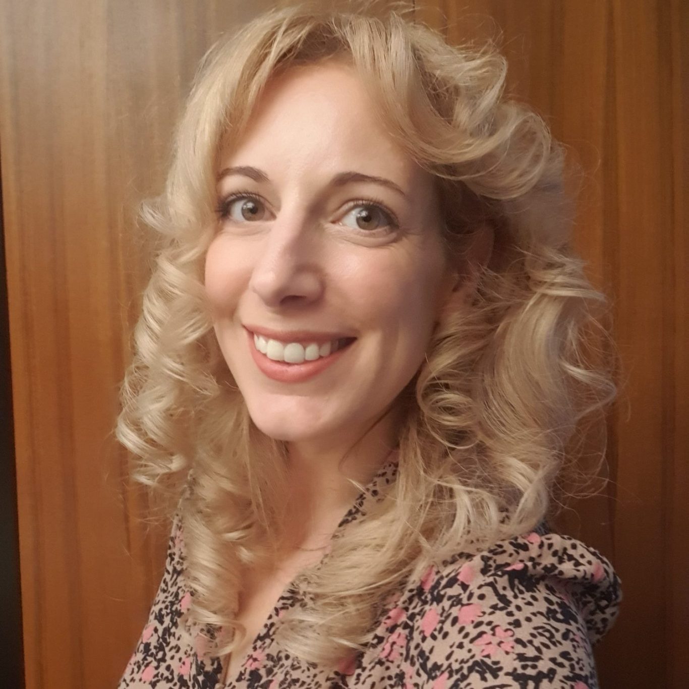 Rebecca, with curly blonde hair smiling, wearing a floral dress against a wooden background.