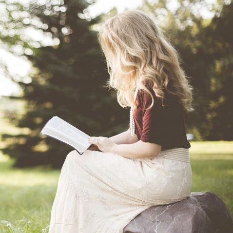 Rebecca sitting on a rock, reading a book in a park with lush greenery.